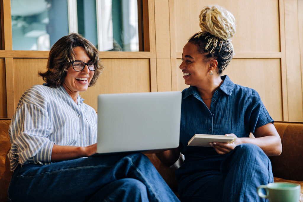 Two adult women enjoy a friendly discussion, using a laptop and a notebook. Their bright smiles convey positivity, teamwork, and productivity in a modern work environment.