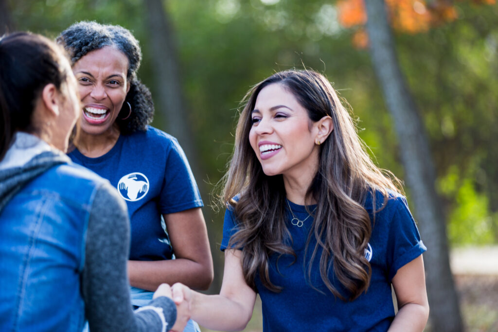 As the mature adult female volunteer smiles, the two mid adult women shake hands and greet each other before the park cleanup begins.